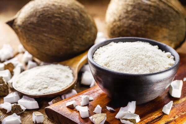 Farinha de coco servida em uma tigela escura, sobre um tábua de madeira, ao lado de uma colher de madeira com mais farinha de coco
