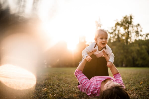 Mãe deitada na grama segura seu bebê ao alto, que dá risada. Ao fundo, árvores e luz solar
