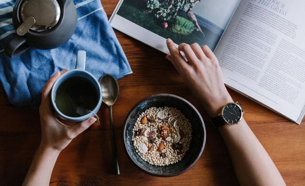 Visão de cima de uma pessoa comendo uma tigela de cereais e chá enquanto lê um livro