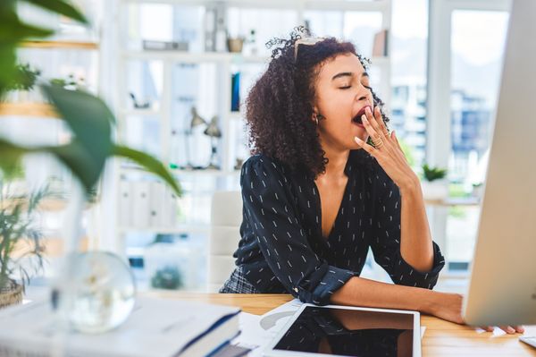 Mulher negra, com cabelos crespos, sentada em frente a um notebook cinza, bocejando