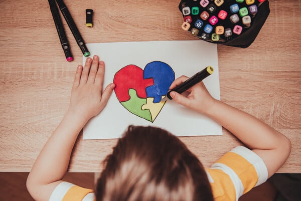 Imagem vista de cima de um garoto loiro e branco, vestindo camiseta amarela e branca listrada, desenhando um quebra-cabeça colorido em formato de coração, em referência ao símbolo do autismo